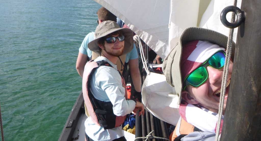 Two people wearing life jackets, hats and sunglasses look at the camera and smile, while they stand on a sailboat